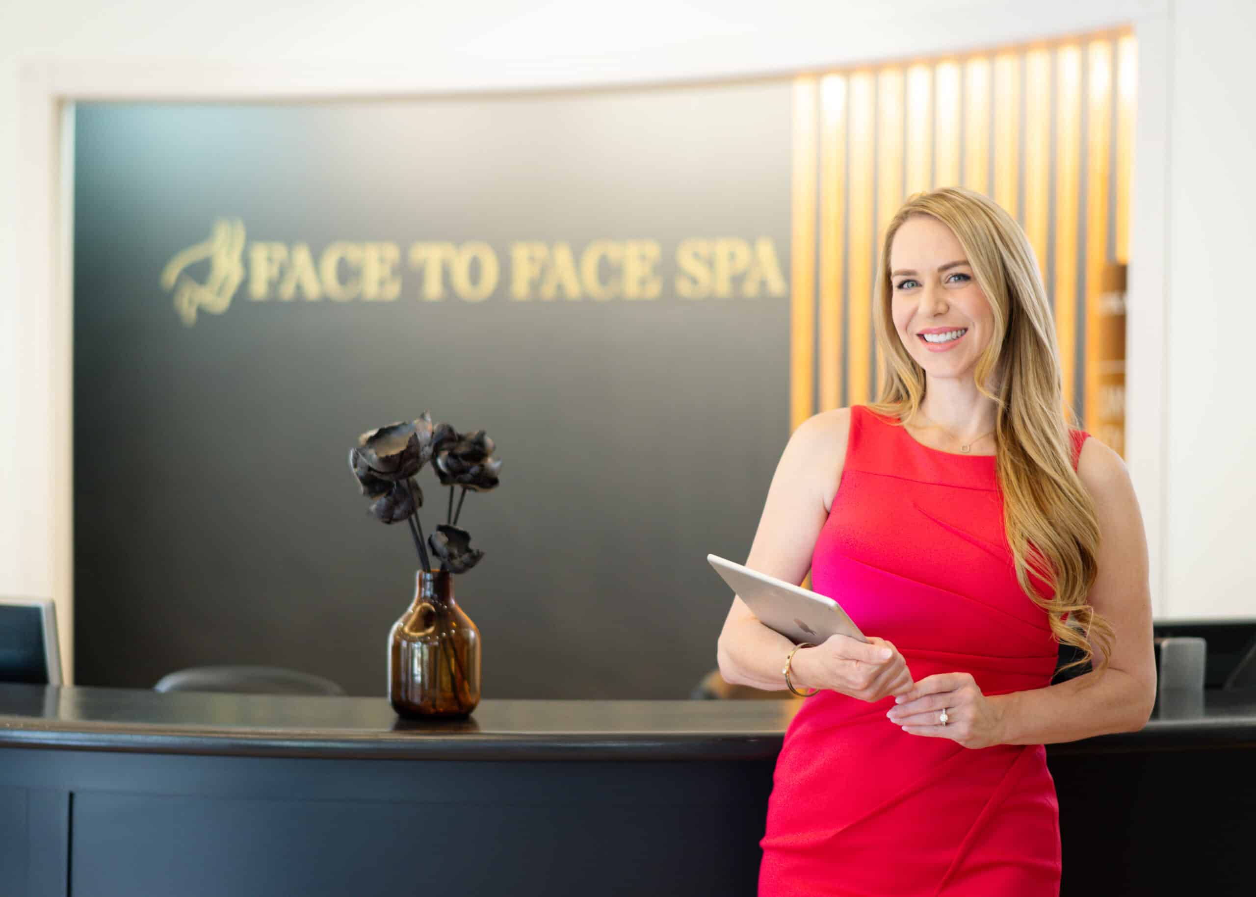 A professional woman in a red dress stands smiling in front of a dark reception desk with a gold "Face to Face Spa" sign behind her.