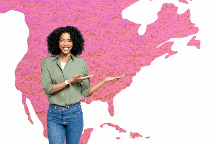 A smiling woman in jeans and a button-down green shirt gestures with her hands towards the pink map of North America that she is standing in front of.