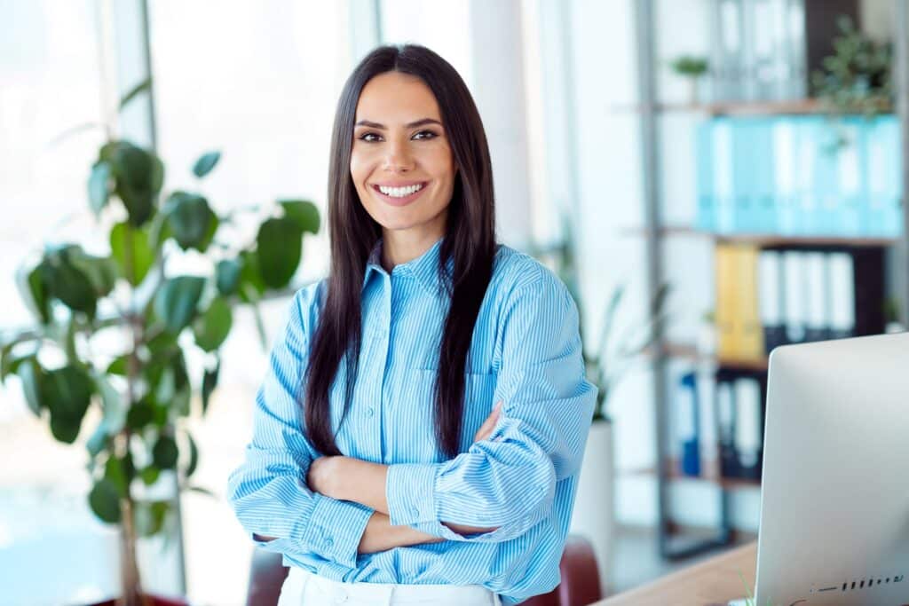 A smiling woman in a button-down blouse with her arms crossed leans against a desk with a computer monitor on it and a fiddleleaf plant and document folders behind her.