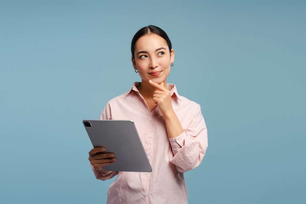A young adult woman in a button-drown dress shirt holding a tablet with one hand and her chin with the other while making a thoughtful expression.