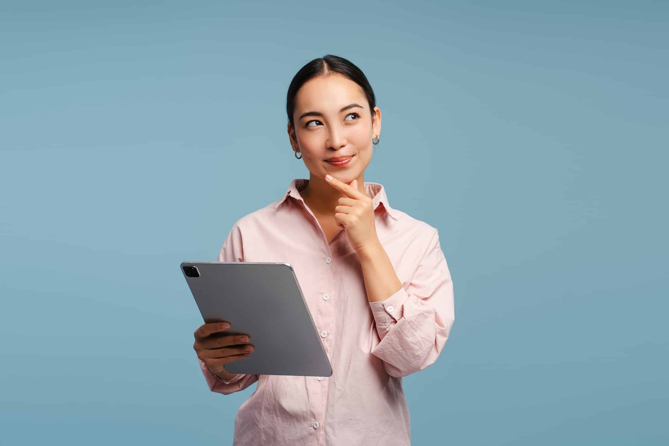 A young adult woman in a button-drown dress shirt holding a tablet with one hand and her chin with the other while making a thoughtful expression.