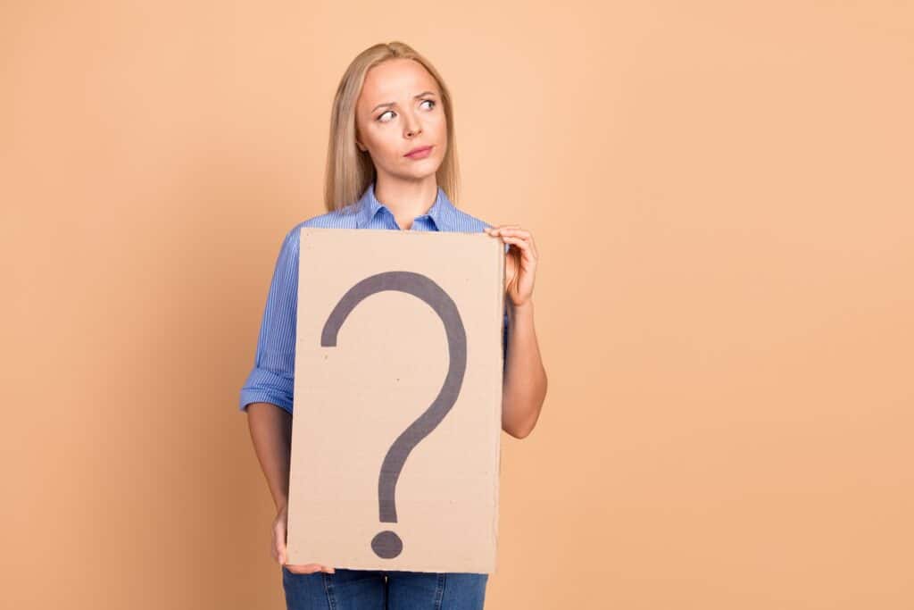 A woman with a thoughtful expression on her face looks to the side while holding a large piece of cardboard with a large black question mark drawn on it.