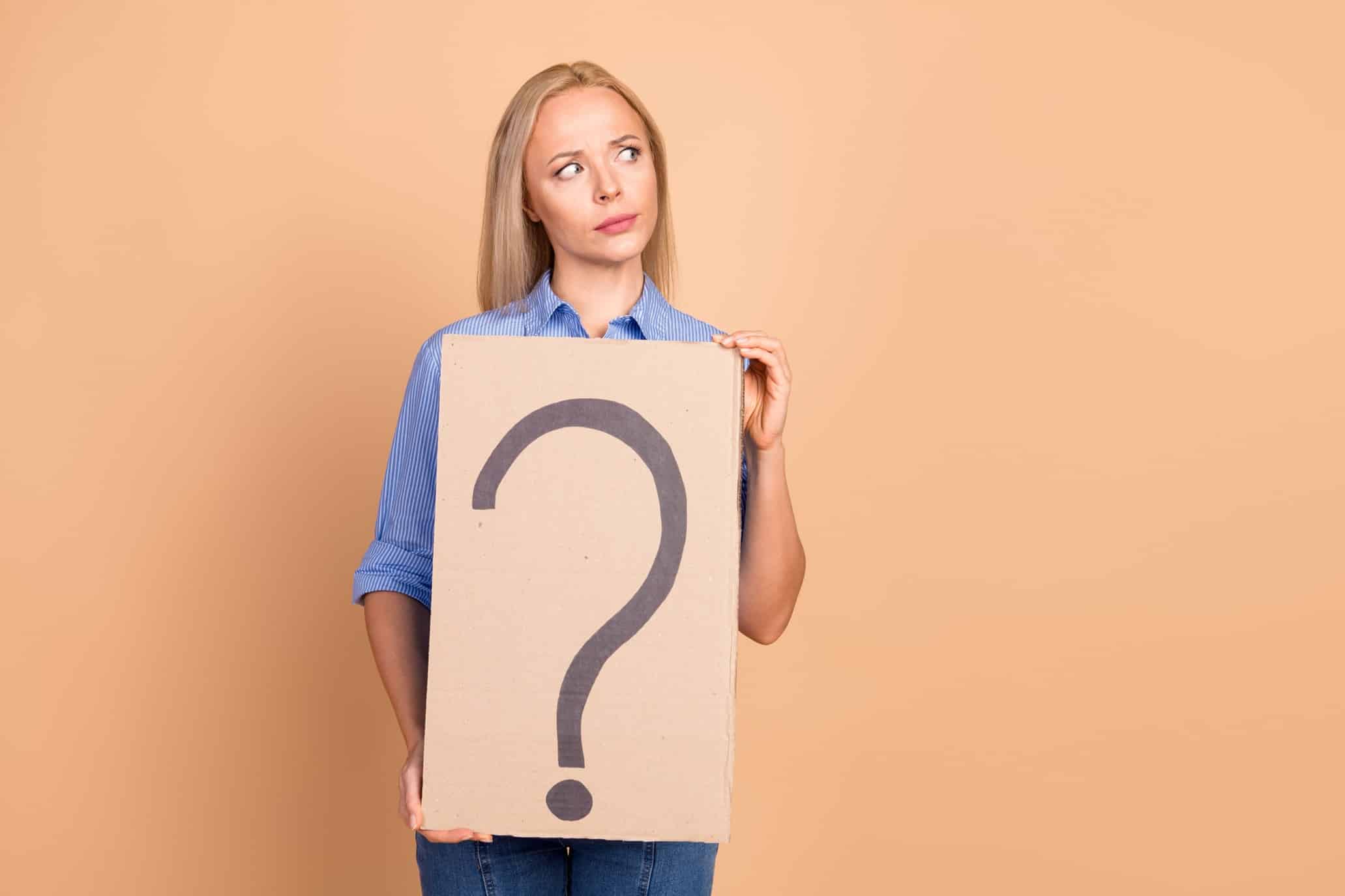 A woman with a thoughtful expression on her face looks to the side while holding a large piece of cardboard with a large black question mark drawn on it.