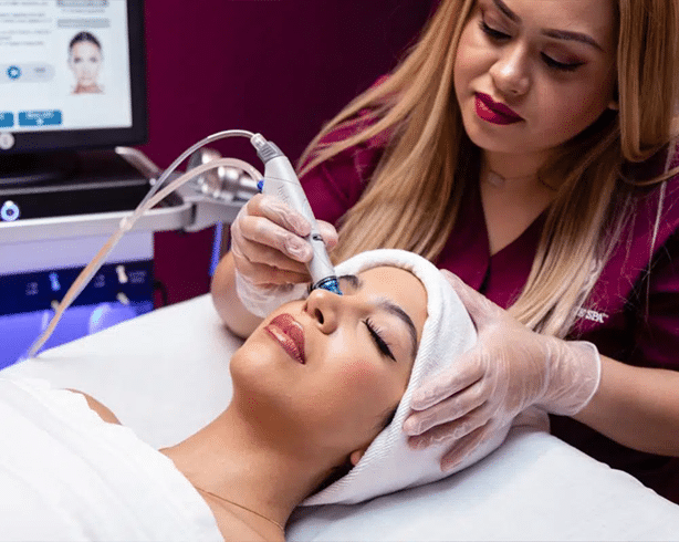 An esthetician uses an electronic skincare tool on the face of a woman lying down on a table with her eyes closed and a towel around her hair.