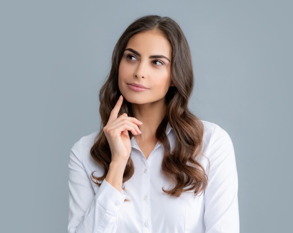 A woman with brown hair holding her hand to her face and looking up with an inquisitive expression.