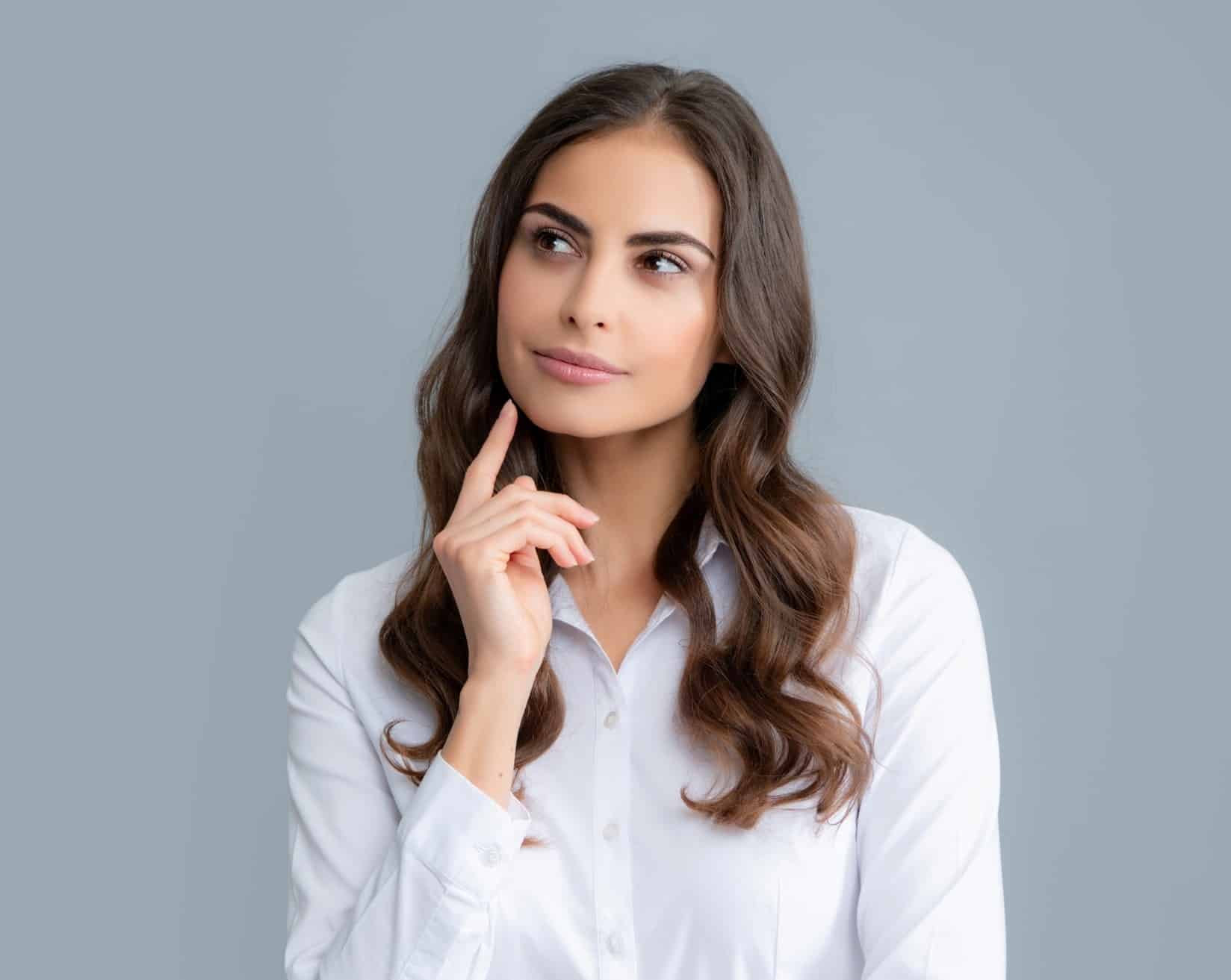 A woman with brown hair holding her hand to her face and looking up with an inquisitive expression.